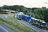 Flere lastebiler står i kø på en rampe ved en truckstop, omgitt av skog og veier.